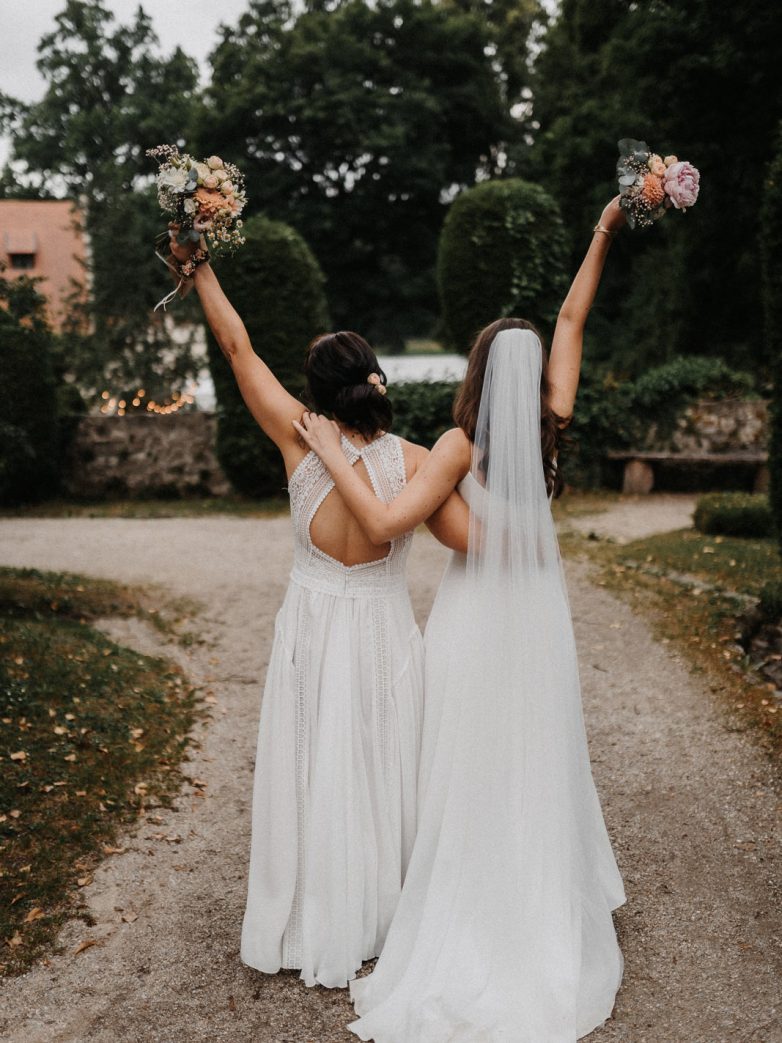 Zwei Frauen in Hochzeitskleidern heben ihre Arme mit Blumensträußen